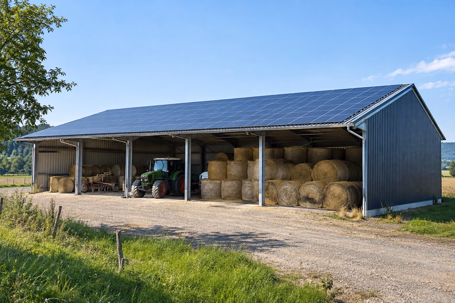 Hangar agricole métallique ouvert avec toiture couverte de panneaux solaires, bottes de paille et tracteur à l’abri